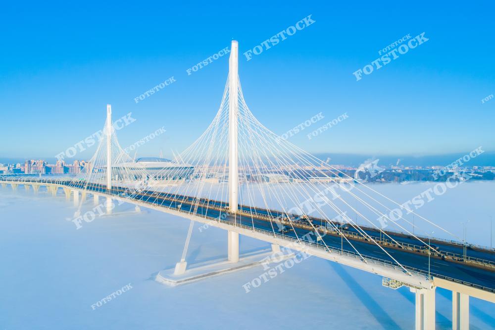 На фотографии изображен Вантовый мост, или Большой Обуховский мост, перекинутый через Финский залив. Белоснежные опоры и ванты моста контрастируют с голубым зимним небом. Под мостом ледяная поверхность залива подчеркивает зимний характер пейзажа. Этот мост — важная часть транспортной инфраструктуры города, соединяющая его районы.